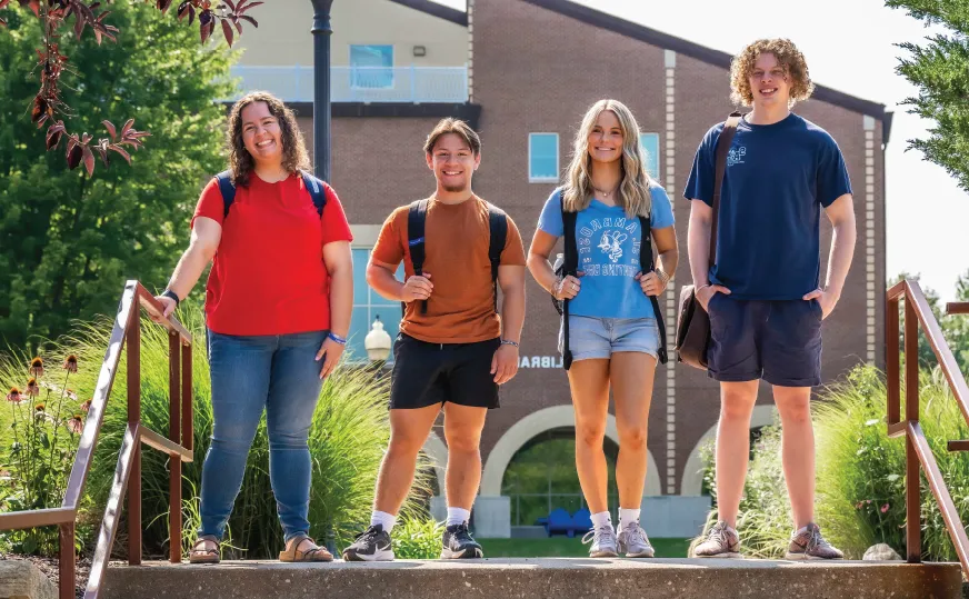 Four students standing outdoors in front of the 分图书馆, smiling.